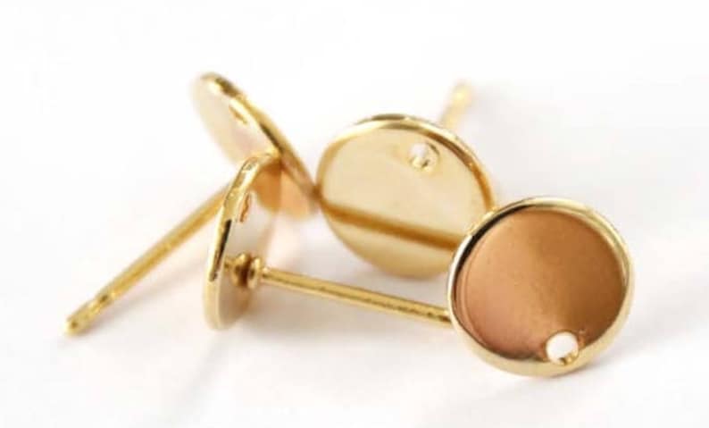 May include: Three gold-toned earring settings with a round, flat base and a post for attaching earrings. The settings have a small hole in the center for inserting a stone or other embellishment.
