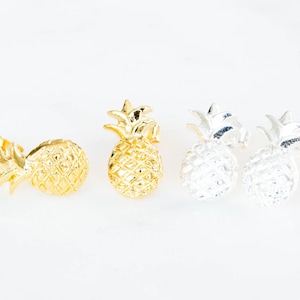 May include: Gold and silver-colored pineapple stud earrings. The earrings are in the shape of a pineapple, with textured surfaces and detailed leaves. The earrings are displayed on a white background.