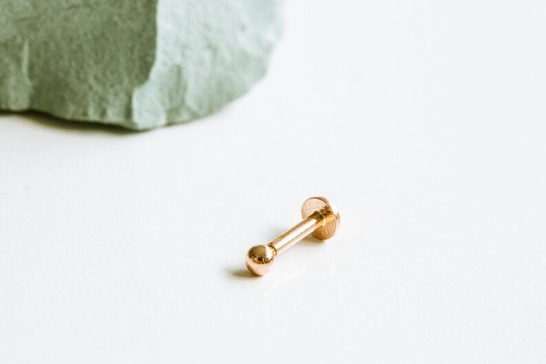May include: A gold-colored nose ring with a small ball on one end and a flat disc on the other end.