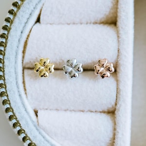 May include: Three clover-shaped stud earrings in gold, silver, and rose gold, displayed in a white jewelry box. The earrings are small and detailed, with a four-leaf clover design. The box has a gold zipper.