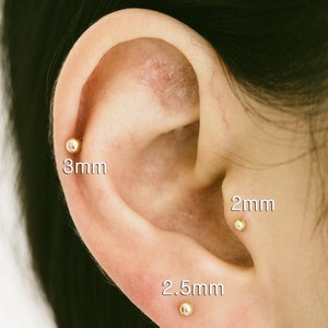 May include: Close-up of a person's ear with three gold ball earrings. The earrings are 2mm, 2.5mm, and 3mm in size.