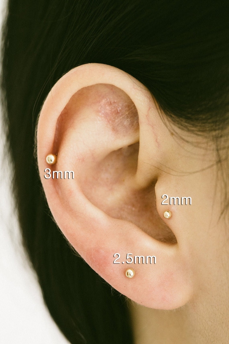 May include: Close-up of a person's ear with three gold ball earrings. The earrings are 2mm, 2.5mm, and 3mm in size.