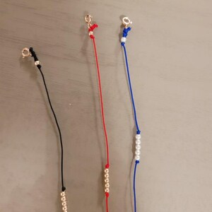 Pu&ograve; includere: Tre braccialetti con cordini e perline di diversi colori. Il primo braccialetto ha un cordino nero e perline dorate. Il secondo braccialetto ha un cordino rosso e perline dorate. Il terzo braccialetto ha un cordino blu e perline bianche.