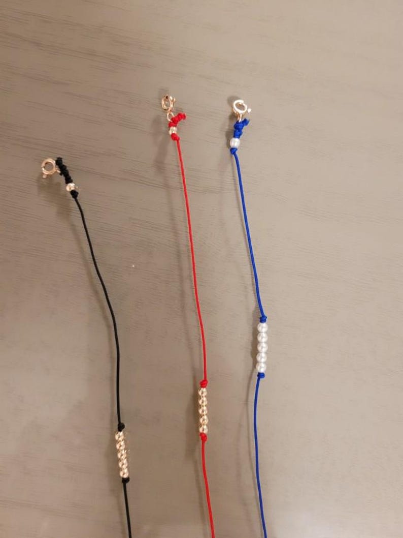 Pu&ograve; includere: Tre braccialetti con cordini e perline di diversi colori. Il primo braccialetto ha un cordino nero e perline dorate. Il secondo braccialetto ha un cordino rosso e perline dorate. Il terzo braccialetto ha un cordino blu e perline bianche.
