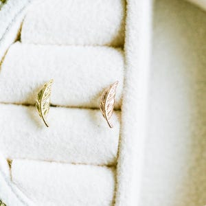 May include: Two feather-shaped stud earrings, one gold and one rose gold, displayed in a white jewelry box. The earrings are detailed with delicate lines, resembling a feather's texture. The box has a gold zipper.