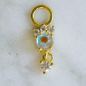 Pode incluir: Um pingente dourado com uma parte superior circular e um design pendurado. O pingente apresenta uma pedra central iridescente cercada por cristais claros e brilhantes. Um grupo de cristais menor está na parte inferior.
