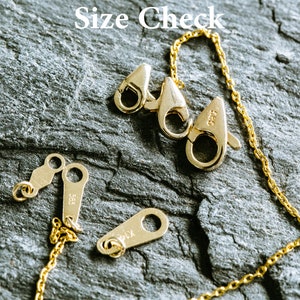 May include: Close-up of a gold chain necklace with a lobster clasp and two jump rings. The chain is on a dark gray textured surface. The text "Size Check" is visible in the top center of the image.