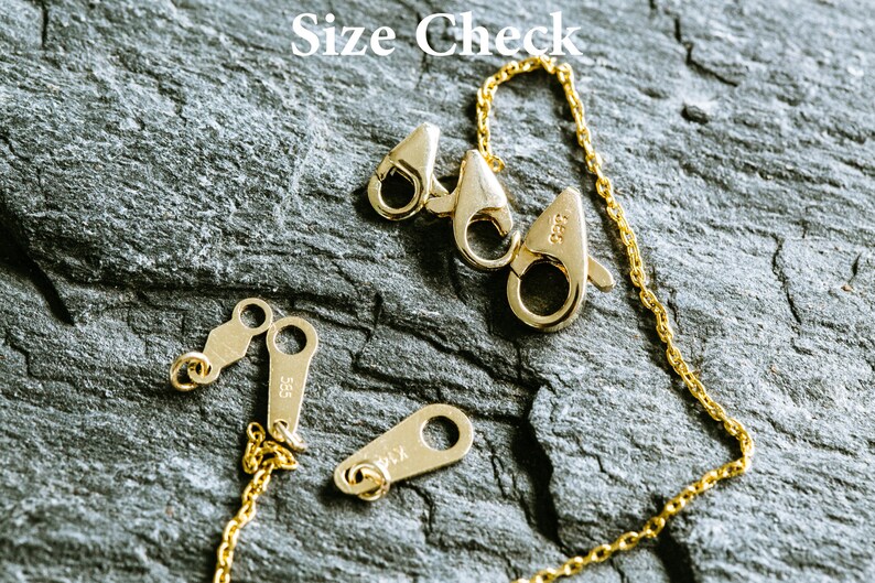 May include: Close-up of a gold chain necklace with a lobster clasp and two jump rings. The chain is on a dark gray textured surface. The text "Size Check" is visible in the top center of the image.