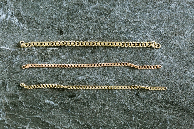 May include: Three gold chains of different lengths and thicknesses laid out on a gray textured surface.