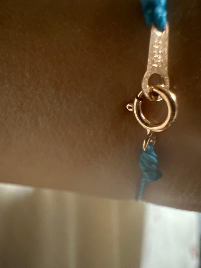 Pu&ograve; includere: Primo piano di un braccialetto in corda blu turchese con una chiusura dorata. La chiusura ha un piccolo anello rotondo e un design decorativo.