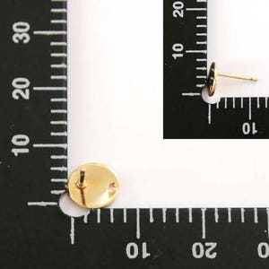 May include: A close-up of a gold-colored earring with a round, flat back. The earring is measured against a ruler.