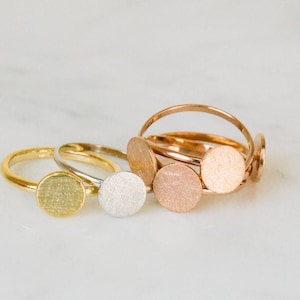 Puede incluir: Tres anillos de oro con tapas circulares texturizadas. Los anillos están apilados uno encima del otro. El anillo superior es de oro, el anillo del medio es de plata y el anillo inferior es de oro rosa.