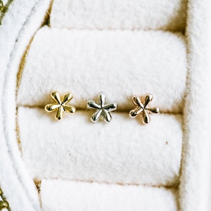May include: Three flower-shaped stud earrings in gold, silver, and rose gold. The earrings are displayed in a white jewelry box with a gold zipper. The earrings are small and delicate, perfect for everyday wear.