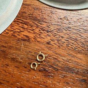 Pu&ograve; includere: Due anelli di salto in metallo dorato, noti anche come chiusure a moschettone, sono su una superficie di legno. Gli anelli sono aperti e pronti per essere attaccati a una catena o altri gioielli.