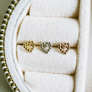 May include: Three heart-shaped stud earrings in gold, silver, and rose gold, each crafted from small, round beads. The earrings are displayed in a white jewelry box with a gold zipper.