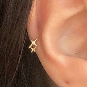 Könnte beinhalten: Ein goldfarbener sternförmiger Ohrring wird am Ohr getragen. Der Ohrring hat ein Doppelstern-Design, wobei ein Stern etwas kleiner ist als der andere. Der Ohrring ist ein kleines, zartes Schmuckstück.