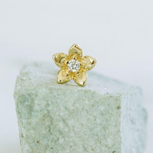 May include: A gold-colored flower-shaped stud earring with a clear, round gemstone center. The earring is set on a light green, textured stone against a white background. The earring's petals are detailed.