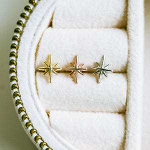 May include: A close-up shot of a white jewelry box with a gold zipper, containing three star-shaped stud earrings. The earrings are gold, rose gold, and silver. The box has a soft, velvety interior.