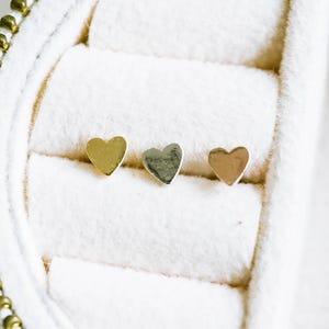 May include: Three heart-shaped stud earrings in gold, silver, and rose gold, displayed in a white jewelry case. The earrings are small and simple, perfect for everyday wear. The case has a gold zipper.