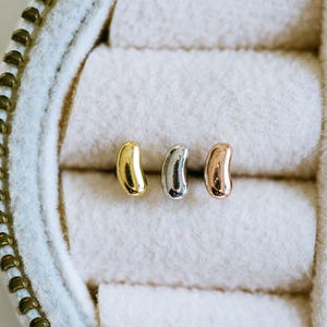 Puede incluir: Tres pendientes de botón con forma de judía en oro, plata y oro rosa. Los pendientes se muestran en un joyero de color claro con un interior suave y texturizado. La caja tiene una cremallera dorada.