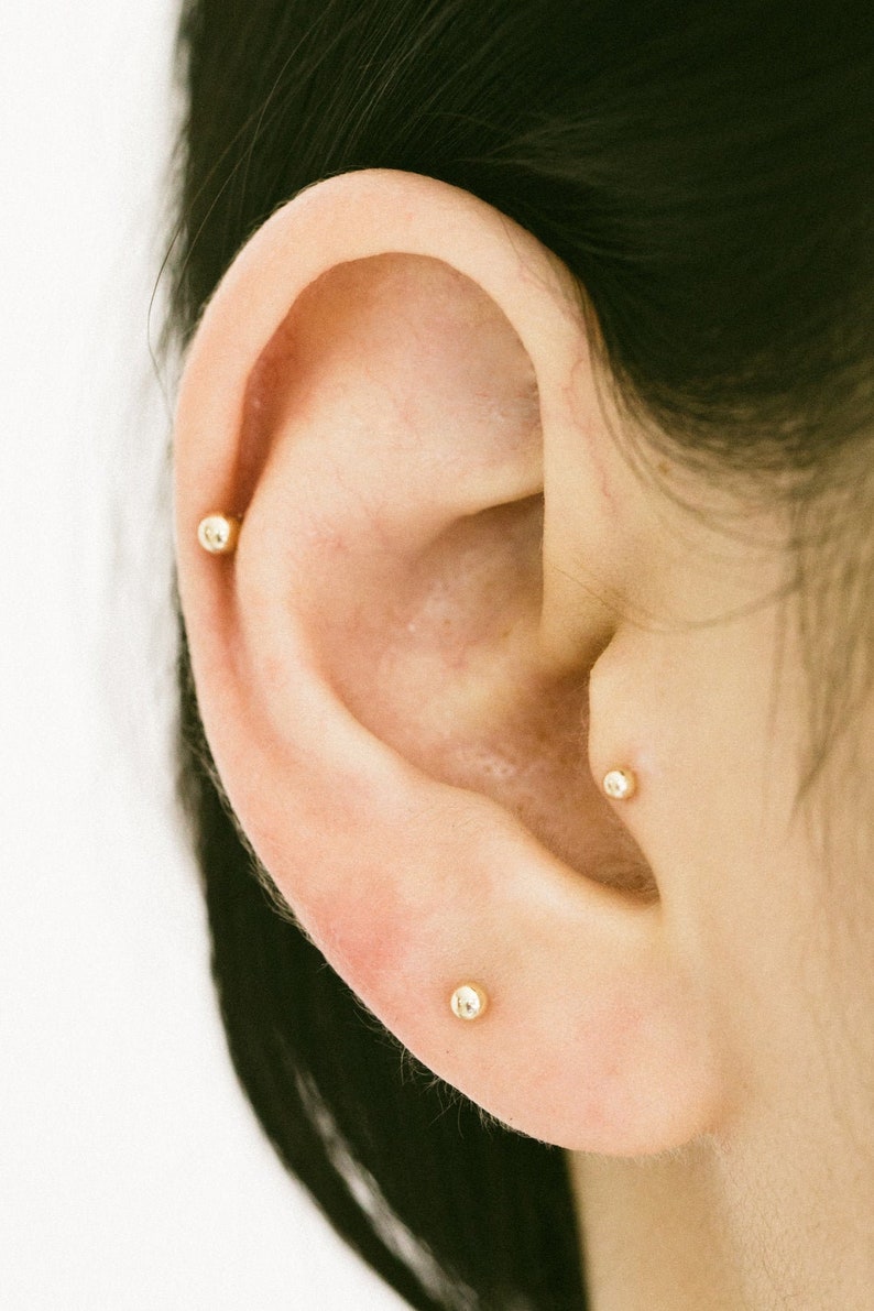 May include: Close-up of a person's ear with three small gold stud earrings. The earrings are in the lobe, helix, and conch piercings.