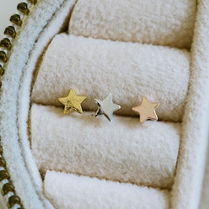 May include: Three small star-shaped stud earrings in gold, silver, and rose gold. The earrings are displayed inside a white jewelry box with a gold zipper. The stars are evenly spaced and have a simple, minimalist design.