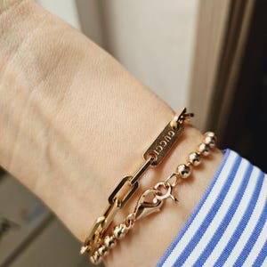 Pu&ograve; includere: Un braccialetto a catena dorato con un'etichetta rettangolare che dice "GUCCI" e un braccialetto di perline dorate.