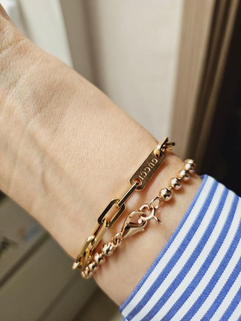 Pu&ograve; includere: Un braccialetto a catena dorato con un'etichetta rettangolare che dice "GUCCI" e un braccialetto di perline dorate.