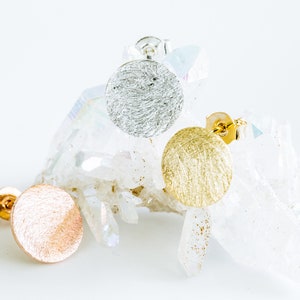 May include: Three pairs of round stud earrings in silver, gold, and rose gold. The earrings are textured and have a brushed finish. The earrings are displayed on a white crystal.