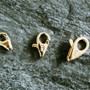 May include: Three gold-toned metal clasps for jewelry making. The clasps are different sizes and have a spring-loaded mechanism. The clasps are marked with the number "585", which indicates they are made of 14 karat gold.