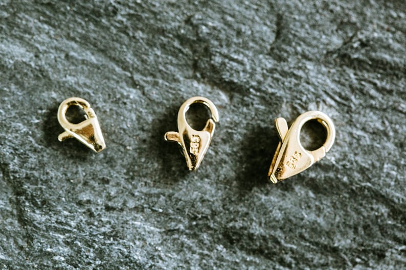 May include: Three gold-toned metal clasps for jewelry making. The clasps are different sizes and have a spring-loaded mechanism. The clasps are marked with the number "585", which indicates they are made of 14 karat gold.