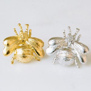 Peut inclure: Deux broches en forme d'abeille, l'une dorée et l'autre argentée, avec des ailes détaillées et un corps texturé.