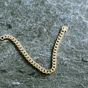May include: A gold chain necklace with a curb link style. The chain is laying on a gray textured surface.