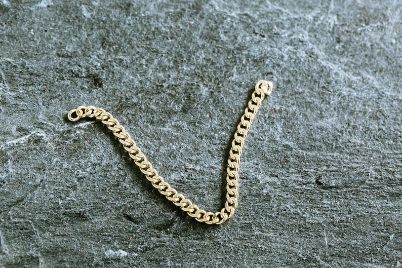 May include: A gold chain necklace with a curb link style. The chain is laying on a gray textured surface.