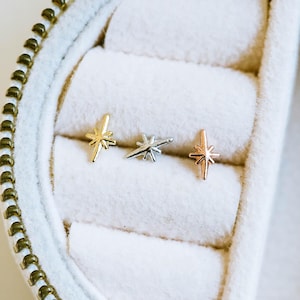Puede incluir: Tres pendientes de estrella en oro, plata y oro rosa se exhiben en una caja de joyería blanca. Los pendientes son pequeños y delicados, con un diseño simple y minimalista. La caja tiene una cremallera dorada.