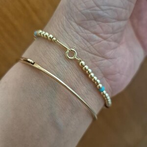 Pu&ograve; includere: Due braccialetti d'oro al polso di una persona. Il braccialetto superiore &egrave; un braccialetto di perline con piccole perline turchesi. Il braccialetto inferiore &egrave; un semplice braccialetto d'oro.