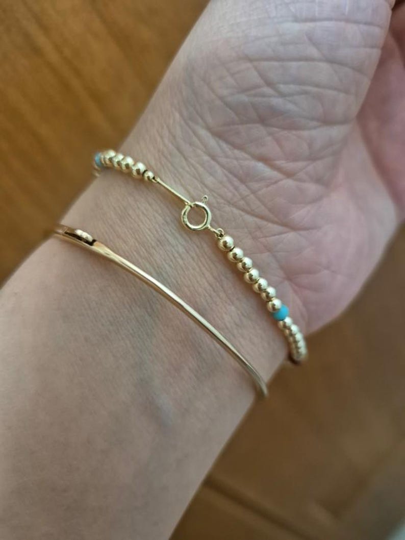 Pu&ograve; includere: Due braccialetti d'oro al polso di una persona. Il braccialetto superiore &egrave; un braccialetto di perline con piccole perline turchesi. Il braccialetto inferiore &egrave; un semplice braccialetto d'oro.