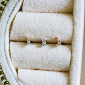 May include: Three crescent moon earrings in gold, silver, and rose gold, each encrusted with small, clear gemstones. The earrings are displayed inside a white, fabric-lined jewelry box with a gold zipper.