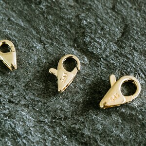 May include: Three gold-toned lobster clasps, each with a different size. The clasps are on a dark gray textured surface.