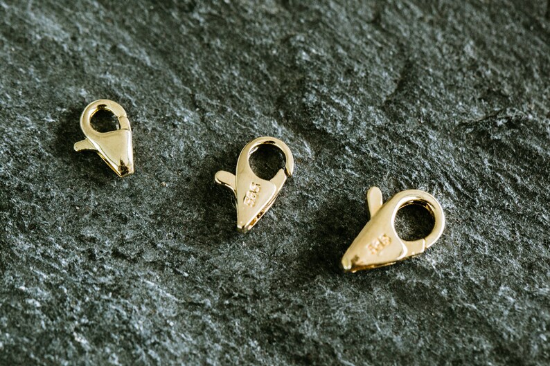 May include: Three gold-toned lobster clasps, each with a different size. The clasps are on a dark gray textured surface.