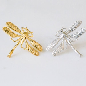 May include: Two dragonfly brooches, one gold and one silver, with intricate details. The brooches are on a white background.
