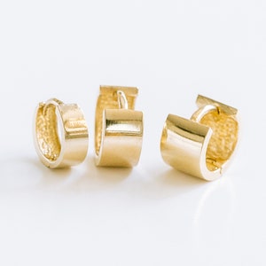 Puede incluir: Tres pendientes de aro de oro, cada uno con un ancho ligeramente diferente. Los pendientes están sobre un fondo blanco.