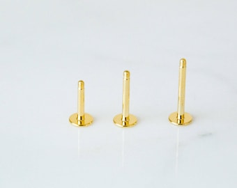 May include: Three gold-coloured body jewellery pieces, each with a flat circular base and a straight post, arranged in order of increasing length.