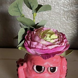 May include: A pink ceramic octopus planter with googly eyes and red beads around the tentacles. A pink and green flower with green leaves sits on top of the planter.
