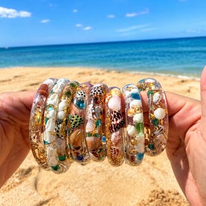 May include: A collection of clear resin bangles embedded with various small seashells, tiny green and blue glass beads, and gold-toned metallic flakes. Each circular bracelet features a unique arrangement of natural white, brown, and spotted shells.