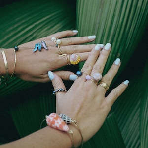 May include: A pair of hands wearing multiple gold rings with various shell and pearl accents. The rings are on a background of green leaves.
