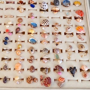 Puede incluir: Una colección de anillos dispuestos en una bandeja organizadora de joyas de color beige. Los anillos presentan diversos elementos naturales, como pequeñas conchas, perlas, piedras de colores y bandas de metal en tonos dorados y plateados.