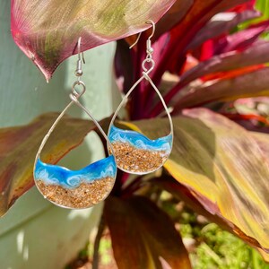 May include: Silver teardrop earrings with a beach scene design. The earrings have a silver wire frame and a clear resin interior. The resin is filled with sand, blue and white resin to mimic ocean waves.