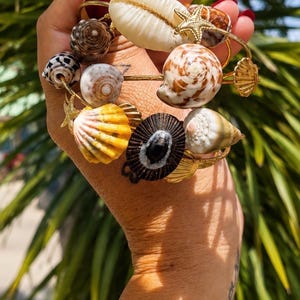 May include: A gold-toned bangle bracelet adorned with a variety of natural seashells, including a large white cowrie, a spiral brown and white shell, a yellow fan shell, and a small gold-toned starfish charm, paired with a wide engraved gold cuff.