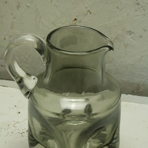 May include: A gray glass pitcher with a rounded body and a curved handle. The pitcher has a spout for easy pouring. The glass has a smoky, translucent appearance, and the design is simple and elegant.